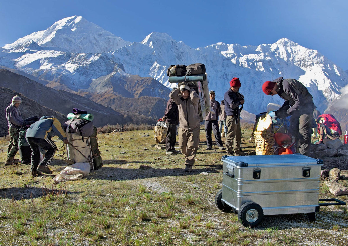 ZARGES K424 XC aluminum case on the off road wheel set (41819) at a Himalaya expedition camp, porters and snow-covered peaks behind